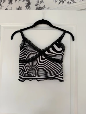 Svartvit ribbad linnetopp med spets, XS - Snygg croppad linnetopp med svartvitt psykedeliskt mönster och ribbad struktur. Toppen har tunna axelband, spetsdetaljer vid urringningen och vågig kant nertill. I storlek XS, från H&M.