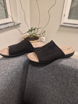 Svarta slip-in sandaler 41 från Din-sko - Säljer ett par svarta slip-in sandaler med bred ovandel i mockaimitation och mjuk, beige innersula. Sandalerna har låg sula och öppen tå, perfekta för en avslappnad och stilren look.