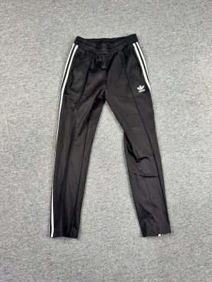 Svarta Adidas mjukisbyxor med vita ränder - Säljer ett par klassiska svarta mjukisbyxor från Adidas med vita ränder längs sidorna och Adidas-logga på vänster ben. Byxorna har resår i midjan och dragkedjefickor. Perfekta för chill eller träning, tillverkade i ett mjukt och följsamt material.