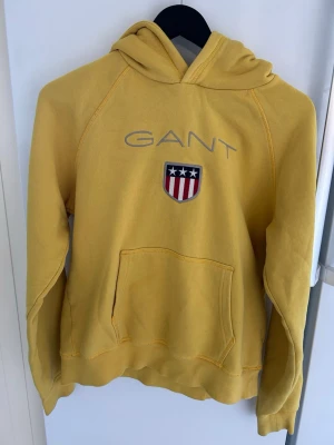 Gul hoodie från GANT med logga - Gul hoodie från GANT med broderad logga och amerikanskinspirerat märke på bröstet. Klassisk känguruficka framtill och huva. Tillverkad i mjuk bomullsblandning, perfekt för en avslappnad och sportig stil.
