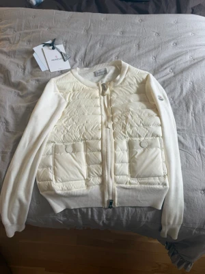 Vit cardiganjacka från Moncler - Snygg vit cardiganjacka från Moncler med quiltad front och två stora fickor med lock. Jackan har ribbade muddar, dragkedja framtill och Moncler-logga på ärmen. Ärmar och rygg är stickade medan framsidan är vadderad för extra värme.