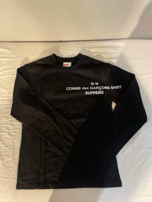 Svart långärmad topp Supreme x Comme des Garçons - Svart långärmad topp från Supreme x Comme des Garçons med vit text på bröstet. Klassisk rund halsringning och rak passform. Materialet är mjuk bomull och designen är enkel men ikonisk med tydlig logga framtill.