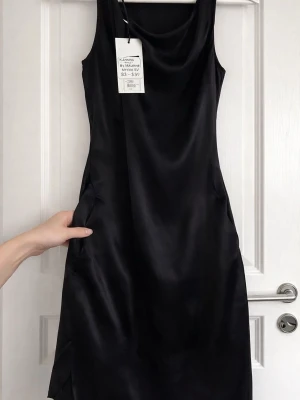 By Malene Birger sidenklänning “Myrime”, strl 38 - Elegant svart sidenklänning i minimalistisk design med fint fall och drapering framtill. Perfekt till fest eller uppklätt tillfälle.  Ny med tags 94% siden med stretch Helfodrad Sidfickor  Diskret innerknäppning (designfunktion) som kan användas eller tas bort vid behov.  Obs: Inneretiketten har släppt lite. (påverkar inte användning).  Nypris ca 2895 kr.