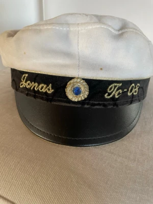 Studentmössa med brodyr och plyschklubba - Studentmössa i vitt tyg med svart skärm, guldbroderad text och blå sten framtill. Medföljer en blå och gul plyschklubba med texten 'STUDENTEN' broderad på framsidan. Perfekt för studentfirande och studentflak.   Stor Jonas på men ksn målas över med tuschpenna eftersom det är svart under  den är använd 1 gång på student   Ny studentpresent dom klubbs ca 45 cm lång 