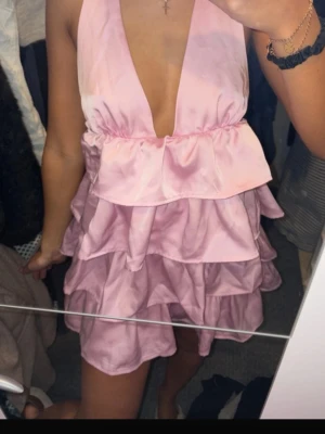 Rosa volangklänning  - Supersöt rosa kortklänning med flera volanger och djup v-ringning. Klänningen har ärmlös design och är gjord i ett glansigt, satinliknande material som ger en lyxig känsla. Perfekt för dig som vill sticka ut med en feminin och trendig look. Från shein