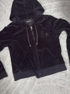 Svart velourhoodie från Juicy Couture - Säljer en svart velourhoodie från Juicy Couture med dragkedja, huva och fickor framtill. Klassisk JC-logga broderad på bröstet och matchande svarta snören med silverfärgade detaljer. Perfekt för en chill och trendig look.