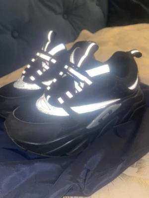 Svarta chunky sneakers med reflexdetaljer - Säljer ett par svarta chunky sneakers från Buffalo med feta sulor och coola reflexdetaljer. Ovandelen är i mesh och syntet, och skorna har en sportig vibe med svarta och vita kontraster. Perfekta för dig som vill sticka ut och gillar streetwear.