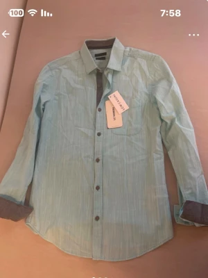 Ljusblå skjorta från Lee Cooper - Snygg ljusblå skjorta från Lee Cooper med diskreta vertikala ränder och klassisk krage. Skjortan har långa ärmar med kontrasttyg på insidan av manschetterna och knappar framtill. Perfekt för en clean och fräsch look.