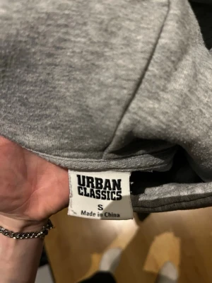 Grå hoodie från Urban Classics - Säljer en enkel och stilren grå hoodie från Urban Classics i storlek S. Hoodien har huva med dragsko och är tillverkad i mjukt material, perfekt för en avslappnad look. Passar dig som gillar basic streetwear och vill ha något bekvämt till vardags.