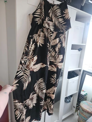Jumpsuit med palmblad - Jumpsuit i svart med beige palmbladsmönster och tunna axelband som knyts upptill. Jumpsuiten har en loose passform och är ärmlös, vilket gör den perfekt för varma dagar. Tillverkad i ett lätt och mjukt material som känns skönt mot huden.