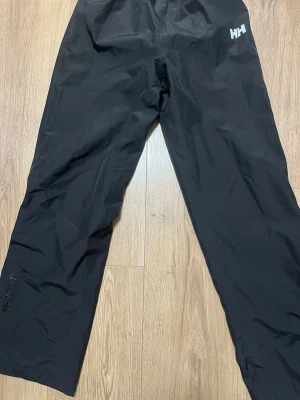 Svarta täckbyxor från Helly Hansen - Svarta täckbyxor från Helly Hansen med elastisk midja och raka ben. Byxorna har diskret logga på ena benet och är tillverkade i ett vattenavvisande material, perfekta för kalla och blöta dagar.