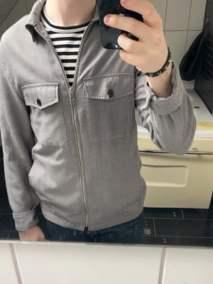 Grå overshirt med dragkedja - Säljer en stilren grå overshirt med dragkedja framtill och två bröstfickor med knappar. Overshirten har klassisk krage och är tillverkad i ett mjukt tyg som känns bekvämt. Perfekt att bära över en t-shirt för en avslappnad look. Från h&m