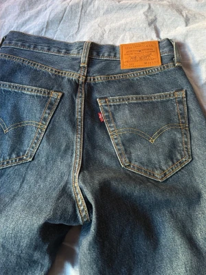 Levi's blå raka jeans - Säljer ett par klassiska blå Levi's jeans med raka ben och hög midja. Jeansen har tydliga kontrastsömmar, två bakfickor med den ikoniska V-formade sömmen och en brun läderpatch bak i midjan. Tillverkade i slitstark denim, perfekta för en avslappnad stil.