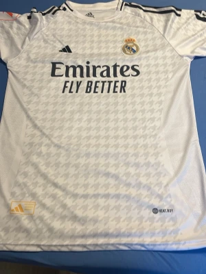Real Madrid matchtröja Adidas vit - Säljer en vit Real Madrid matchtröja från Adidas med svarta detaljer och klubbmärke på bröstet. Tröjan har trycket 'Emirates Fly Better' och är tillverkad i ett lätt, tekniskt material med HEAT.RDY-teknologi. Snyggt mönster över hela framsidan och klassiska Adidas-ränder på axlarna.