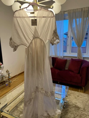 Vitt nattlinne med spets från Aéryne - Långt vitt nattlinne från Aéryne med romantiska spetsdetaljer vid ärmslut och nederkant. Nattlinnet har v-ringning, lös passform och korta ärmar i ett skirt, mjukt material som känns lyxigt mot huden.