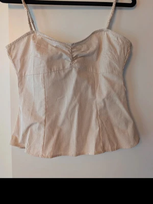 Beige och vit randig linnetopp - Snygg linnetopp med tunna axelband i beige och vitt randigt mönster. Toppen har en lätt rynkning vid bysten och är tillverkad i ett mjukt, svalt material som passar perfekt till varma dagar. Enkel och stilren design som är lätt att matcha.