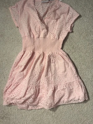 Ljusrosa kort klänning med broderi - ALDRIG ANVÄND MED PRISLAPP KVAR!! ÄR STORLEK 152 MEN ÄR VÄLDIGT STOR I STORLEKEN! Söt kort klänning med broderade blommönster och volangkant. Klänningen har omlott-ringning, korta ärmar och smock i midjan för en bekväm passform. Jättefin till sommaren och t.ex. midsommar! Säljer eftersom jag aldrig hade användning till den eftersom jag fick den på hösten! Den är väldigt stor i storleken och passar mig som är 165 och storlek S! Kostar 600 från början!! Köpte på kids brand store! EJ GENOMSKINLIG