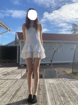 Vit kort klänning med volang - Söt vit kort klänning med smockad överdel och volangig kjol. Klänningen är ärmlös och har broderade spetsdetaljer på kjolen. Perfekt för varma dagar och somriga tillfällen.