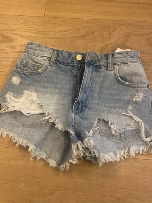 Ljusa denimshorts med fransar - Säljer ett par ljusblå denimshorts med slitna detaljer och fransig kant. Shortsen har fem fickor, bälteshällor och knappstängning. Perfekta för sommaren och ger en avslappnad vibe. Aldrig använda. 