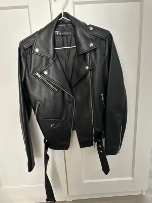 Svart skinnjacka från Zara - Snygg svart skinnjacka från Zara med klassisk bikerstil, silvriga dragkedjor och knappar. Jackan har breda slag, flera fickor med dragkedja och ett bälte i midjan för extra attityd. Perfekt för dig som vill ha en edgy look.