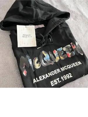 Svart hoodie Alexander McQueen - Snygg svart hoodie från Alexander McQueen med färgglad logga och texten 'EST. 1992' på bröstet. Hoodien har huva med dragsko och en stor magficka. Materialet ser ut att vara mjukt bomullstyg, perfekt för en avslappnad streetwear-look.