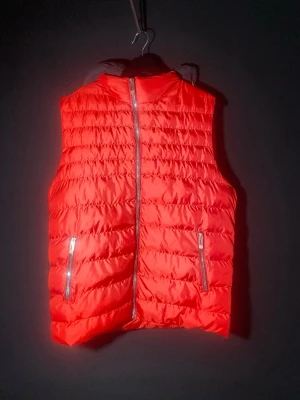 Röd dunväst med dragkedjor - Snygg röd dunväst med quiltad design och två praktiska fickor med dragkedja framtill. Västen har hög krage och stängs med en silverfärgad dragkedja. Perfekt för lager-på-lager och ger en sportig vibe till din outfit.