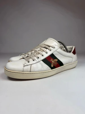 Gucci Ace vita sneakers med bi - Gucci Ace sneakers i vitt läder och gröna och röda sidoband samt ett bi på sidobandet. Skorna har låga skaft, vita snören och kontrasterande hälflikar i rött och grönt.😁