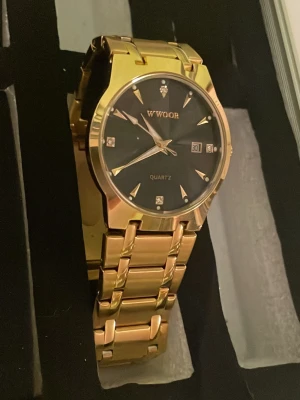 Guldfärgad klocka från WWOOR - Stilren guldfärgad klocka från WWOOR med svart urtavla och datumvisning. Klockan har en klassisk länkarmband i metall och markerade index med små stenar för extra detalj. Perfekt accessoar för dig som vill sticka ut.