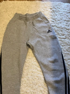 Gråa mjukisbyxor från Adidas - Snygga grå mjukisbyxor från Adidas med svarta ränder längs sidorna och klassisk logga på låret. Byxorna har resår i midjan och sidofickor. Perfekta för chill eller träning, tillverkade i mjuk bomullsblandning.