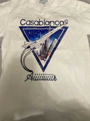 Vit Casablan t-shirt med tryck - Säljer en vit t-shirt från Casablanca med stort grafiskt tryck framtill som visar ett flygplan och stadssilhuett i blått och lila. T-shirten har rund hals och korta ärmar. Perfekt för dig som gillar statement-plagg och coola prints.