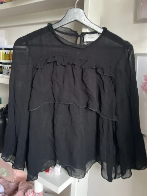 Svart volangblus från Neo Noir - Snygg svart blus från Neo Noir i ett tunt, transparent material. Blusen har volangdetaljer framtill och vid ärmslut samt rund halsringning med knapp i nacken. Perfekt för dig som gillar en luftig och trendig look.