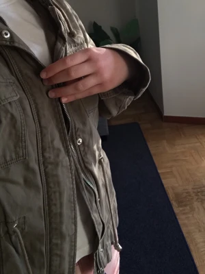 Olivgrön jeansjacka med luva  - Snygg olivgrön jeansjacka med luva och dragsko i midjan. Jackan har flera fickor framtill, dragkedja och tryckknappar. Perfekt för blåsiga dagar och enkel att matcha med olika outfits.