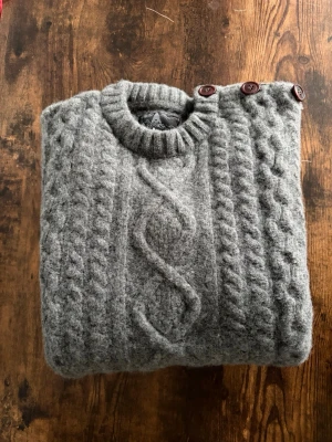 Barbour Sweatshirt - Barbour stickad tröja i storlek L. Klassisk kabelstickad design i grå färg med knappdetaljer vid axeln. Väldigt clean old money/casual vibe. Bra skick utan större defekter. Perfekt till jeans eller chinos för en stilren outfit.