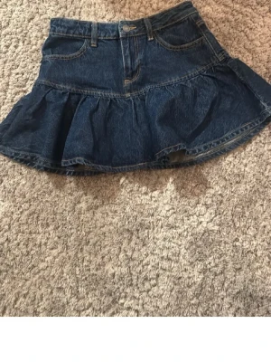 Mörkblå jeansskort med volang - Säljer en snygg mörkblå jeansskort med volangkant och klassiska fickor fram. Skorten har en feminin siluett och är gjord i denim med synliga sömmar. Perfekt för dig som gillar en avslappnad men trendig stil.