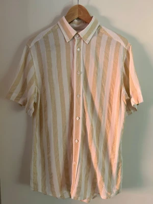 Kortarmad linneskjorta från These Glory Days - Kortärmad skjorta med breda beige och vita ränder. Klassisk krage och knäppning framtill med vita knappar. Tillverkad i ett lätt och luftigt material, perfekt för varmare dagar. Skjortan har en avslappnad passform och är enkel att styla. Priset går att diskutera.