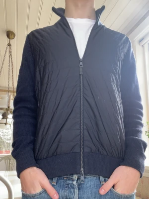 J.lindeberg cardigan  - Säljer en snygg svart jacka från J.Lindeberg i storlek M. Jackan har en quiltad design med dragkedja och en metall logga på ärmen. Perfekt för höst och vår🍁Den är bekväm och i fint skick🌟Byten av rockar och dunjackor är intressant🙏🏼