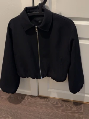 Svart croppad jacka  - Stilren svart bomberjacka med croppad passform och silverfärgad dragkedja framtill. Jackan har klassisk krage, långa ärmar och diskreta sidofickor. Storlek L för en mer oversized fit, är en XS/S i vanliga fall.