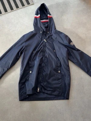 Mörkblå vindjacka från Moncler - Snygg mörkblå vindjacka från Moncler med huva och dragsko. Jackan har en diskret logga på ärmen, två framfickor med tryckknappar och dragkedja framtill. Materialet är glansigt och lätt, perfekt för blåsiga dagar. Röd och vit rand på huvan ger en sportig touch.