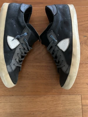 Philippe Model - Snygga mörkblåa sneakers med vit sula och blåa skosnören. Skorna har en låg profil och en vit detalj på sidan. Ovandelen är i mocka och sulan är i gummi. Perfekta för en avslappnad streetstil. Storlek 42 men passar 43. Sulan är sönder men allt annat är bra.