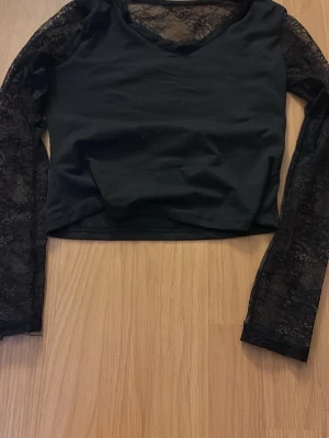 Svart topp med spetsdetaljer - Snygg svart topp med långa spetsärmar och spets vid halsringningen. Modellen är croppad och har en v-ringad urringning framtill. Perfekt för dig som gillar en stilren men ändå lite edgy look.