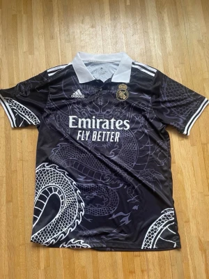 Real Madrid svart matchtröja drake - Säljer en svart Real Madrid matchtröja från Adidas med vit krage och vita detaljer. Tröjan har ett stort drakmotiv i grått över hela plagget, klubbmärke och Adidas-logga på bröstet samt 'Emirates Fly Better' tryckt framtill. Perfekt för dig som älskar fotboll och unik design.