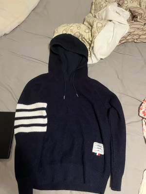 Mörkblå hoodie - Snygg mörkblå hoodiemed klassiska vita ränder på ena ärmen och en patch med märkets logga nedtill. Hoodien har dragsko i huvan och är stickad i ett mjukt material som känns lyxigt. Perfekt för dig som gillar streetwear med exklusiv känsla.