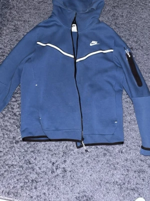 Blå Nike hoodie med dragkedja - Snygg blå hoodie från Nike med vit logga på bröstet och vita detaljer längs bröstet. Tröjan har hel dragkedja, huva och en ficka med dragkedja på ärmen. Tillverkad i mjukt material, perfekt för en sportig och avslappnad stil.