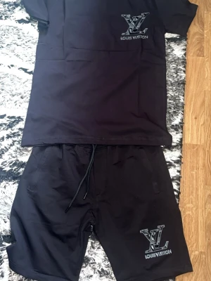 Svart Louis Vuitton set med logga - Snyggt svart set från Louis Vuitton med matchande t-shirt och shorts. Båda plaggen har den ikoniska LV-loggan tryckt i vitt och grått framtill. T-shirten har rund hals och korta ärmar, shortsen har dragsko i midjan och sidofickor. Perfekt för en avslappnad och trendig look.