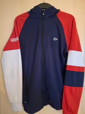 Blå, röd och vit hoodie från Lacoste - Snygg hoodie från Lacoste i blått med röda och vita detaljer på ärmarna. Dragkedja framtill, huva och klassisk Lacoste-logga på bröstet. Materialet känns mjukt och sportigt, perfekt för en avslappnad stil. LACOSTE-märke på ena ärmen.