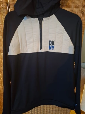 Svart och beige hoodie från DKNY - Snygg svart hoodie från DKNY med beige panel över bröstet och halv dragkedja framtill. DKNY-logga broderad i svart och blått på vänster sida. Hoodien har huva och långa ärmar, perfekt för en avslappnad streetwear-look.