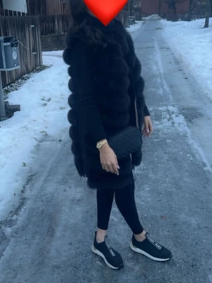 Svart lång pälsväst i fuskpäls - Säljer en svart lång pälsväst i fuskpäls med voluminösa paneler och öppen front. Västen har en modern och trendig look som passar perfekt till lager-på-lager-stil. Enkel att matcha med olika outfits för en cool vibe.
