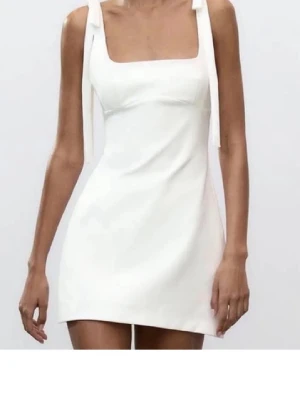 Vit kort klänning med knytband - Vit klänning i en kortare längd med axelband knutna i rosetter där jag har sytt upp axelbanden cirka 5 cm med en tunntråd som enkelt går att sprätta upp. Klänningen har en fyrkantig urringning och har används en gång.