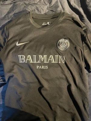 PSG x Balmain Mbappé matchtröja - Snygg svart PSG matchtröja i samarbete med Balmain, med Paris Saint-Germain-logga och Nike-märke på bröstet. Framsidan har Balmain Paris-tryck och baksidan har Mbappé och nummer 7 i vitt. Tillverkad i lätt och ventilerande Dri-FIT-material.