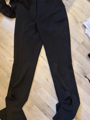 Svarta kostymbyxor från RYVLS - Stilrena svarta kostymbyxor från RYVLS med raka ben och klassisk passform. Byxorna har hällor för skärp och stängs med knapp och dragkedja. Perfekta för en clean och snygg look. Materialet är mjukt och följsamt.